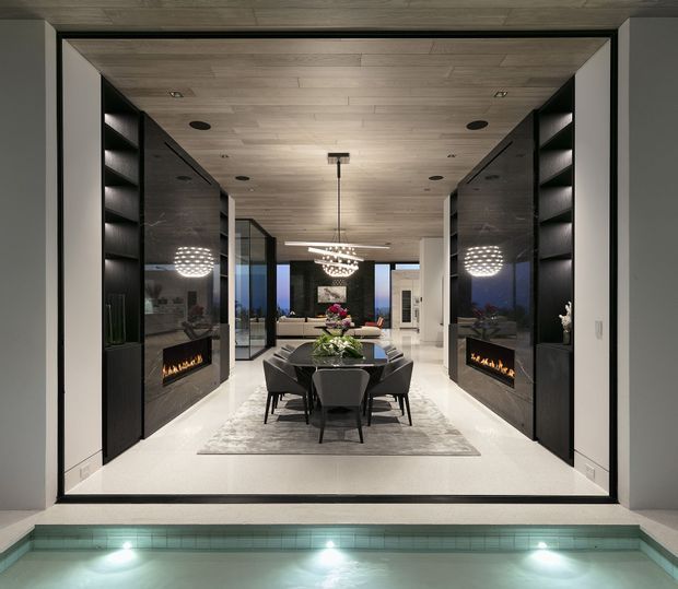 A refined, symmetrical contemporary dining and living area viewed from beside a swimming pool. The room features a dark dining table with upholstered chairs, mirrored black built-in cabinetry incorporating linear fireplaces on both sides, illuminated shelving, a pale rug, extensive glazing, and a warm timber-lined ceiling. Sculptural pendant lights and reflective surfaces create a luxurious evening atmosphere.