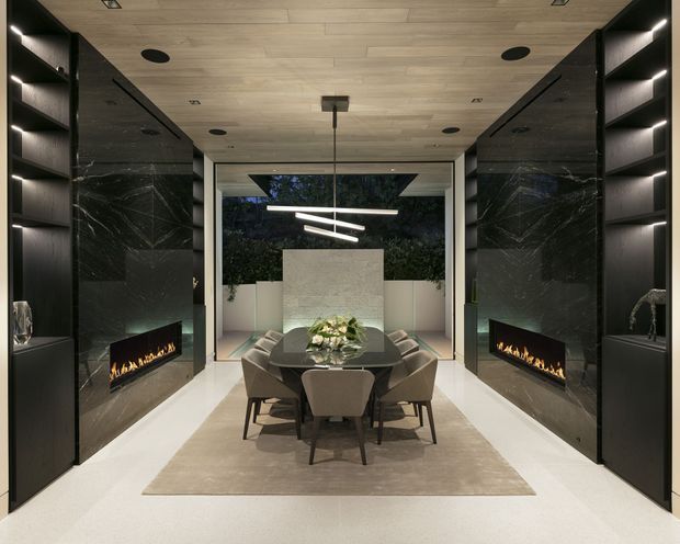 A sophisticated contemporary dining room arranged symmetrically between two dark marble feature walls with integrated linear fireplaces and illuminated display shelving. A central dining table with upholstered chairs sits on a neutral rug, beneath sculptural linear pendant lighting. The room features a light timber ceiling, pale flooring, large glazed opening to an outdoor courtyard, and a refined dark-and-light palette.
