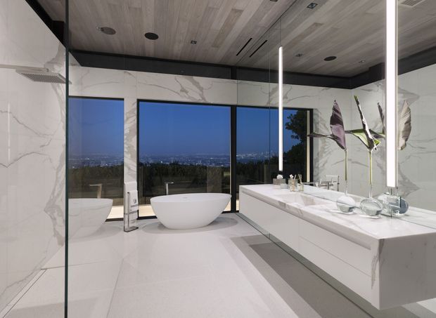 Luxurious contemporary bathroom with a freestanding white bathtub positioned beside floor-to-ceiling glazing, a large double vanity with integrated basins, full-height marble-look wall surfaces, and pale large-format floor tiles. A timber-look ceiling with recessed lighting contrasts with dark ceiling trim and black window frames. The room overlooks a distant cityscape and landscaped hillside.