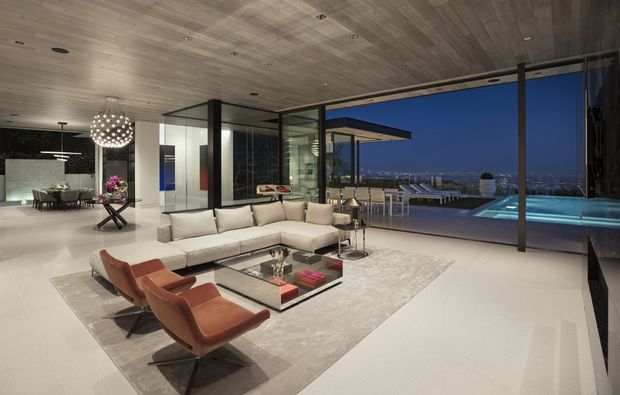 A sophisticated open-plan living room at dusk, featuring a large cream sectional sofa, rust-coloured lounge chairs, a glass coffee table, and a sculptural pendant light. Full-height glazing opens the room to a terrace with a swimming pool and expansive city or waterfront views. The space has a dramatic dark timber ceiling and broad pale flooring, with a sleek contemporary fit-out and strong indoor-outdoor connection.