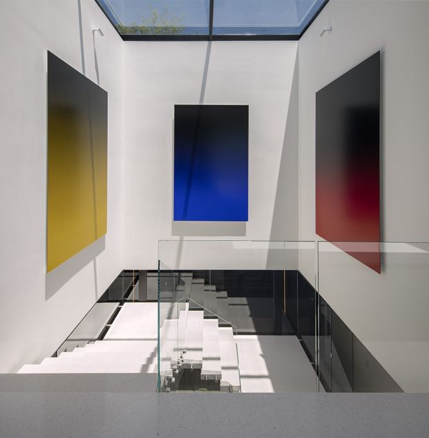 A bright, double-height contemporary stairwell or landing with a central glass skylight, white walls, a sculptural white staircase, and transparent glass balustrades. Large abstract blue, yellow, and red wall panels provide strong colour accents, while sunlight casts geometric shadows across the space.