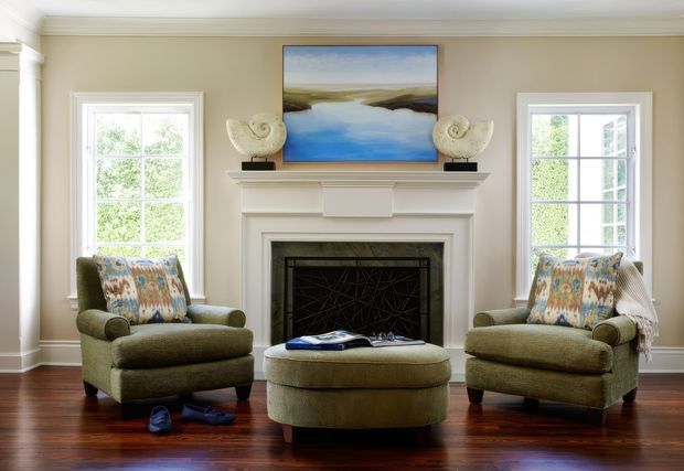 Symmetrical, traditionally styled living room with a central white-painted fireplace and mantel, framed by two tall multi-pane windows. Two olive-green upholstered armchairs and a matching round ottoman sit on dark timber flooring, with a landscape artwork and decorative shell sculptures above the fireplace.