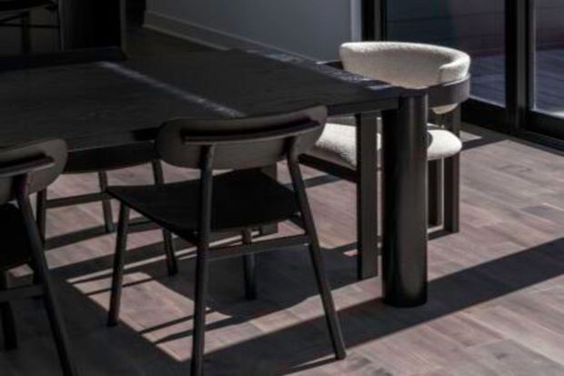 Close-up view of a contemporary dining area featuring a dark rectangular dining table with rounded-edge detailing, several dark-framed upholstered chairs, and a cylindrical pedestal support. The space has dark timber-look flooring and expansive black-framed glazing, with strong sunlight casting geometric shadows across the floor.