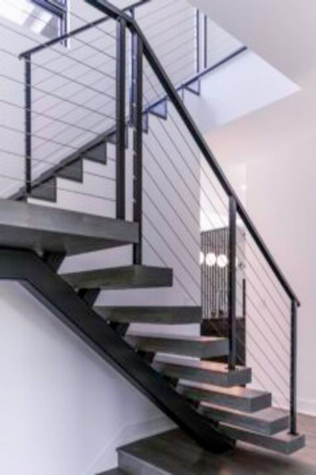 Interior view of a contemporary open-riser staircase with dark structural stringers, thick rectangular treads, slender black metal posts, and horizontal cable balustrading. The stair rises beside a white wall toward an upper landing, with bright natural light from a large rooflight or skylight and a partial glimpse of an adjacent upper-level space.