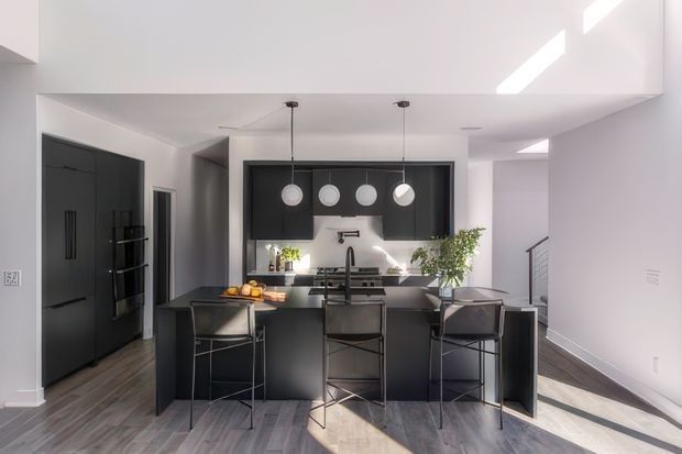 Bright contemporary kitchen with a large black island and three bar stools, dark full-height refrigerator cabinetry, black upper and lower joinery, a white backsplash, and four spherical pendant lights. Grey-toned flooring, white walls and ceiling, and strong natural light create a crisp, high-contrast interior. A stairway is visible at the right.