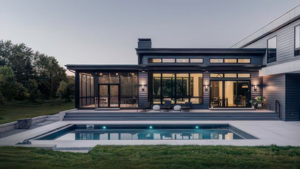 A contemporary multi-level residence viewed from the backyard, with dark horizontal cladding, extensive black-framed glazing, a covered outdoor entertaining area, broad steps, and a rectangular swimming pool in the foreground. The composition highlights the connection between the home and landscaped outdoor space at dusk.
