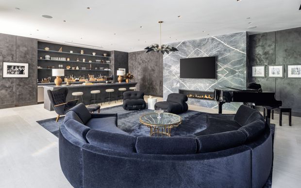 A luxurious contemporary media and entertaining room with a large curved navy sectional sofa arranged around a glass coffee table. Dark textured wall panels, a marble-look fireplace feature wall with linear firebox, wall-mounted television, built-in bar shelving, and a black grand piano create a dramatic, refined interior. Recessed ceiling lighting and pale flooring provide contrast to the dark furnishings and finishes.