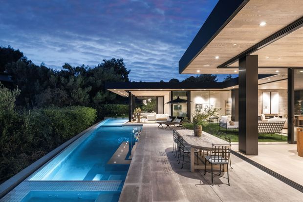 Evening exterior view of a contemporary, flat-roofed residence with extensive open-plan glazing, broad roof overhangs, a covered outdoor dining terrace, and a long illuminated swimming pool integrated into the landscaped setting. Warm interior lighting contrasts with the cool blue dusk and surrounding vegetation.