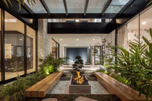 A sophisticated indoor courtyard or atrium viewed from within a glazed residential interior. Timber bench seating lines both sides of a gravel garden with lush planting, stepping stones, and a central square fire feature. Black-framed glazing and a translucent roof create a dramatic indoor-outdoor connection, while a calm living area is visible beyond the courtyard. The design combines warm timber, dark metal framing, stone-like surfaces, greenery, and firelight.
