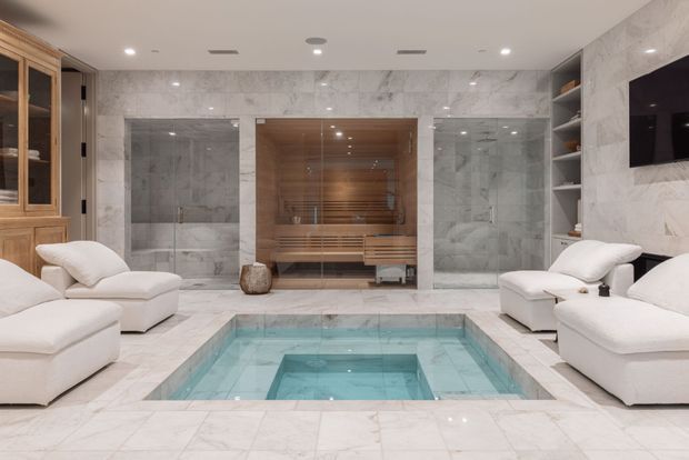 Luxurious indoor wellness area featuring a sunken rectangular plunge pool or spa in the foreground, surrounded by pale marble flooring and walls. A timber-lined sauna with glass doors is centered at the rear, flanked by glazed shower enclosures. White upholstered lounge chairs, built-in timber cabinetry, open shelving, and a wall-mounted television complete the refined spa-like interior.