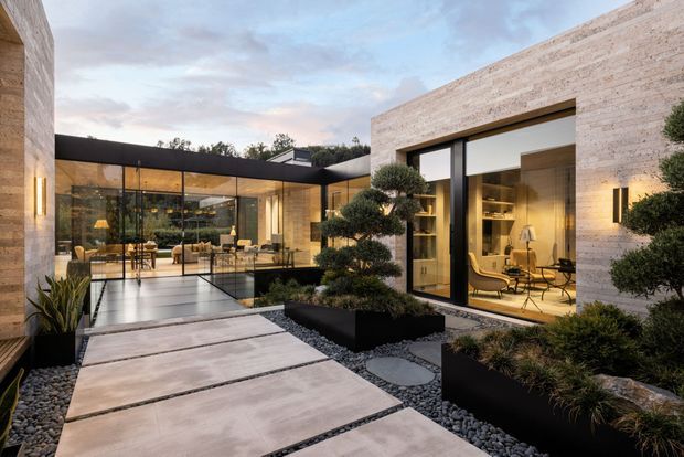 Contemporary courtyard view of a low-profile residence with pale stone-clad walls, extensive floor-to-ceiling glazing, and a dark flat-roof structure. Large rectangular concrete pavers cross a minimalist Japanese-inspired gravel garden with sculpted shrubs and raised black planters. Warm interior lighting reveals a dining and living area on the left and a refined workspace or sitting area on the right.