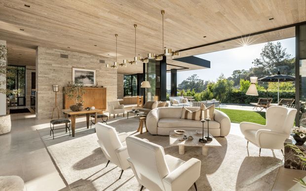 A spacious, refined open-plan living room with a warm timber-lined ceiling, pale stone feature walls, polished concrete flooring, and extensive floor-to-ceiling glazing opening onto a landscaped lawn and patio. The room is furnished with cream sofas, sculptural white lounge chairs, a light coffee table, a timber sideboard, and elegant pendant lighting. A covered outdoor entertaining area and garden are visible beyond the glazing.