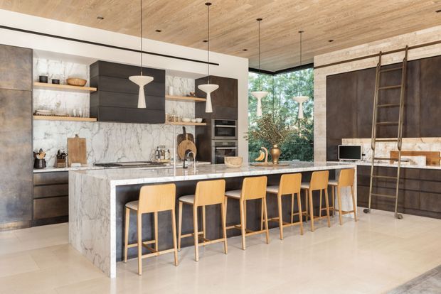 A spacious, high-end open-plan kitchen dominated by a large marble island with seating for six. Dark timber cabinetry contrasts with pale marble surfaces, while a timber-lined ceiling, pendant lights, integrated appliances, open shelving, and a large glazed opening with greenery beyond create a warm contemporary interior. A tall built-in storage wall with ladder is visible on the right.