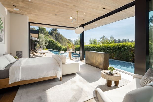 Bright, airy contemporary bedroom with a low timber bed, white bedding, a sculptural pendant light, and a white upholstered lounge chair. Full-height black-framed glazing opens directly to a swimming pool and landscaped outdoor area, while a warm timber-lined ceiling and pale carpet create a calm, refined interior.