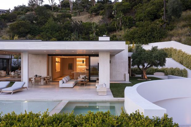 A refined modern home set into a landscaped hillside, viewed from the pool area. The flat-roofed white pavilion has expansive floor-to-ceiling glazing, an open living area with neutral furnishings, a timber-toned kitchen or joinery zone, and a broad paved terrace. A swimming pool and sculptural curved white wall dominate the foreground, with layered garden planting surrounding the property.