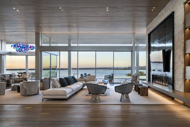 A spacious, refined contemporary living room with expansive floor-to-ceiling glazing overlooking a body of water. The room features warm timber wall and ceiling finishes, a large neutral sectional sofa, sculptural lounge chairs, a built-in linear fireplace within a light stone feature wall, integrated media cabinetry, and recessed lighting. A dining area and colourful pendant light are visible to the left.
