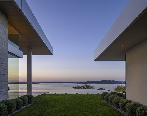 A contemporary waterfront residential exterior viewed from a landscaped lawn courtyard. Two minimalist building wings frame a wide outlook across calm water toward a distant hilly shoreline. The architecture features strong horizontal roof overhangs, floor-to-ceiling glazing, pale stone wall cladding, smooth rendered walls, and timber-toned soffits, photographed at dusk.