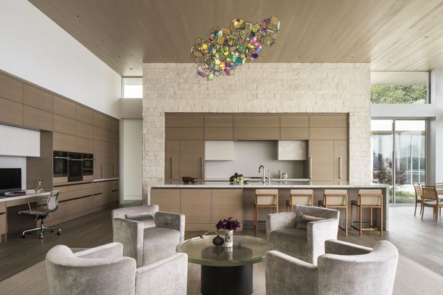 Open-plan contemporary kitchen and living area with extensive warm timber cabinetry, a large island with bar seating, and a lounge zone furnished with curved light-grey armchairs around a round coffee table. A home office nook is integrated into the cabinetry at left. The room features a white textured stone feature wall, timber-lined ceiling, large glazed openings, and a colourful sculptural pendant light.