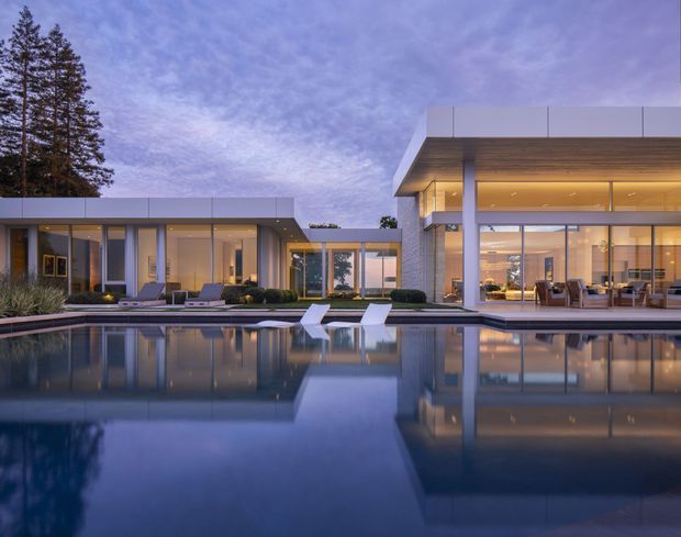 A dusk exterior view of a large contemporary, flat-roofed residence arranged around a reflecting swimming pool. The building has crisp rectilinear volumes, extensive floor-to-ceiling glazing, pale masonry or concrete walls, slim dark window frames, and a deep overhanging roof with a warm timber-lined soffit. The illuminated interiors reveal a dining and living area, while landscaped planting and tall trees frame the property.