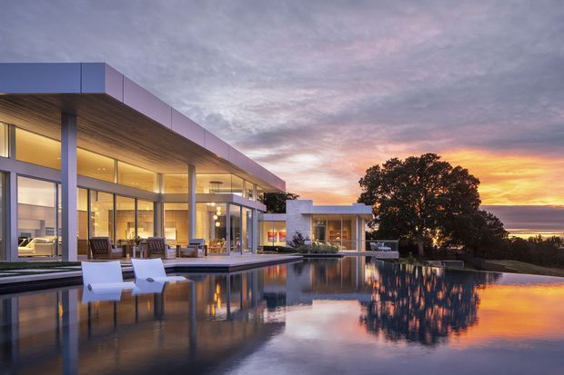 A contemporary flat-roofed luxury residence photographed at sunset, with extensive floor-to-ceiling glazing, deep roof overhangs, slender structural columns, and warm timber soffits. The home opens onto a large reflecting or swimming pool with in-water loungers, while a mature tree and distant coastal or water horizon form the background. The scene has a calm, refined, resort-like atmosphere with strong sunset reflections across the pool.