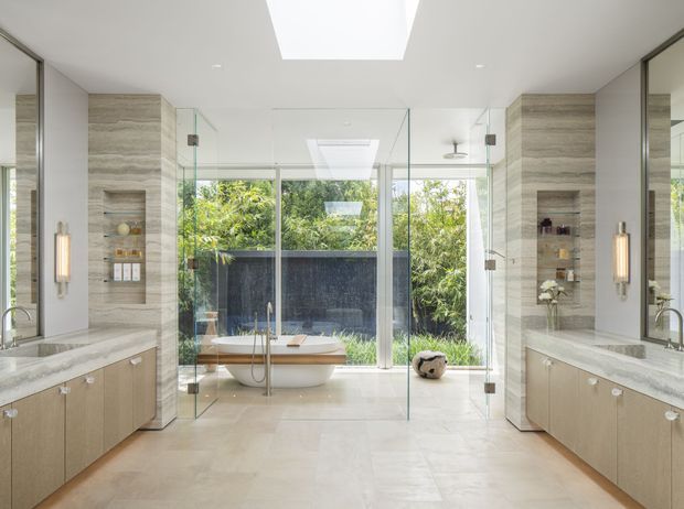 A bright, spacious contemporary bathroom with a freestanding oval bathtub positioned in front of full-height glazing and a lush garden view. Two symmetrical floating vanities with pale wood cabinetry and light stone benchtops flank the room, while glass shower screens, mirrored side walls, recessed ceiling lighting, skylights, and a central vertical stone-clad feature wall create a refined, spa-like interior.