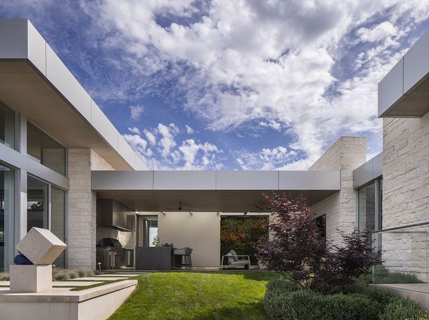 Contemporary low-profile courtyard residence viewed from the landscaped garden. The home features crisp white and pale stone walls, broad floor-to-ceiling glazing, projecting flat-roofed volumes, and a covered outdoor living area enclosing the courtyard. A lawn, clipped planting, and a geometric outdoor sculpture occupy the foreground beneath a dramatic cloud-filled sky.
