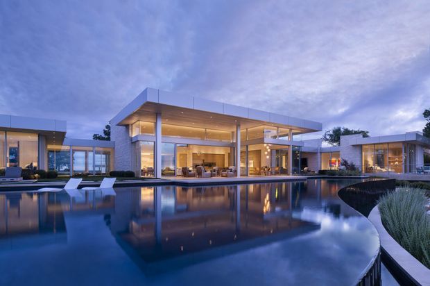 A wide dusk exterior view of a luxurious modernist residence arranged around a large reflecting pool. The home features crisp flat rooflines, extensive floor-to-ceiling glazing, pale stone or rendered walls, slender structural columns, warm recessed lighting, and multiple connected pavilion-like volumes. Poolside loungers and landscaped planting sit in the foreground, while the illuminated living spaces are visible through the glazing.