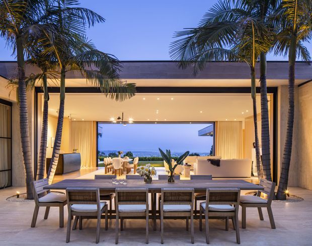 Twilight view of a luxurious open-plan indoor-outdoor living and dining area. A long timber dining table with upholstered chairs occupies the foreground beneath a covered terrace, while a fully open glazed wall connects to a softly furnished living room and reveals a distant ocean or coastal horizon. Tall palm trees frame the space, with warm recessed lighting, timber-lined walls and ceiling, and a clean contemporary pavilion-like design.