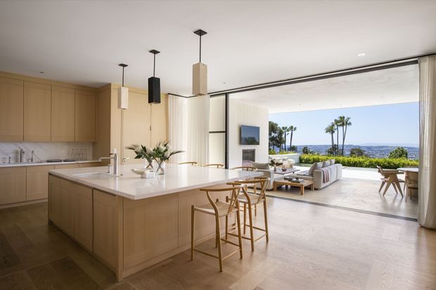 Open-plan kitchen and living area with a large pale timber island, white stone benchtop, integrated light-wood cabinetry, and pendant lights. Floor-to-ceiling sliding glass doors open to a bright terrace and expansive coastal/ocean outlook. The living area contains neutral upholstered seating and a low timber coffee table, creating an airy, refined indoor-outdoor setting.