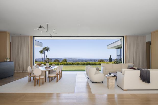 Bright, open-plan contemporary living and dining room with floor-to-ceiling glazing and a wide view toward a landscaped lawn, palm trees, and distant coastal or urban scenery. The room has pale timber flooring, white sofas and sculptural lounge chairs, a light dining table with upholstered seating, full-height beige curtains, and minimalist ceiling lighting. A large television is visible at the left, while the glazing opens visually toward an outdoor terrace or garden.