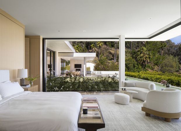 A bright, contemporary bedroom with a large upholstered bed, light timber wall panelling, and full-height black-framed glazing. Curved cream lounge chairs and an ottoman sit beside the window, overlooking a lush landscaped garden and a modern outdoor living area with a fireplace. The room has a calm, refined, indoor-outdoor character with recessed ceiling lighting and a pale textured carpet.