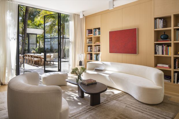 A bright, refined living room with a sculptural cream sofa and matching rounded armchairs arranged around a dark oval coffee table. Full-height black-framed glazing opens to a lush tropical courtyard or terrace, while a warm timber wall incorporates built-in bookshelves and displays. A red abstract artwork, soft curtains, and a large neutral rug complete the calm contemporary interior.