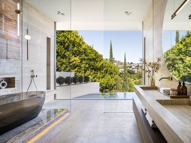Luxurious contemporary bathroom with a freestanding sculptural bathtub, large-format stone or marble wall and floor surfaces, frameless glass shower enclosure, and extensive mirrored or glazed views toward a leafy hillside landscape. A long floating vanity with integrated basin and warm wood-toned cabinetry occupies the right side, while slim brass-toned fixtures and pendant lighting add refined detail. The room is bright, spacious, and strongly connected to the outdoors.