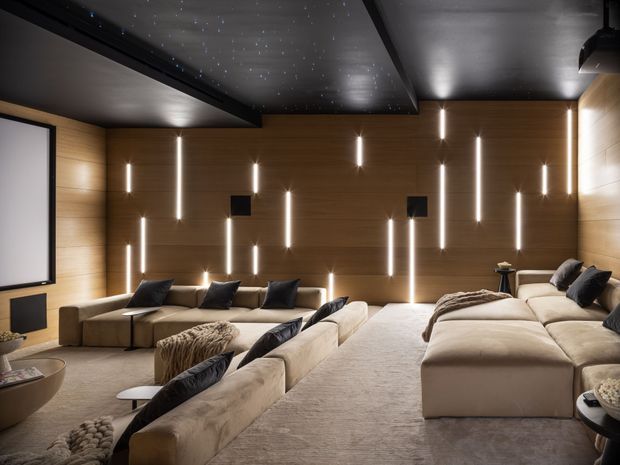 A dark, luxurious home cinema with multiple rows of low, cream-coloured sectional seating facing a large projection screen. Warm timber wall panelling is fitted with numerous vertical linear lights, while the dark ceiling includes recessed star-like pin lights and a projecting beam. Soft neutral carpeting, cushions, throws, and side tables create a refined, immersive screening-room atmosphere.