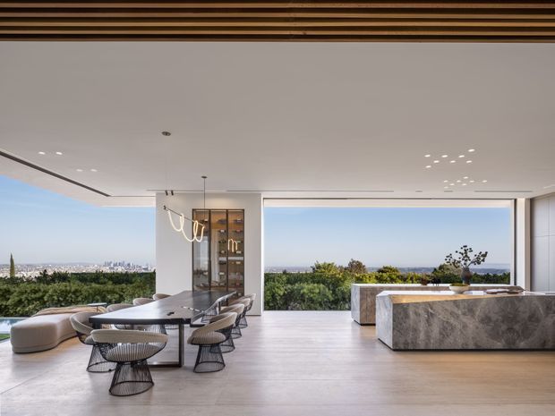 A spacious, high-end open-plan interior combining a dining area and kitchen, with expansive floor-to-ceiling glazing overlooking a landscaped setting and distant city views. The room has pale timber flooring, white walls and ceiling, a dramatic stone kitchen island, dark-framed dining furniture, a sculptural pendant light, and a linear timber ceiling feature. The space feels bright, refined, and contemporary.