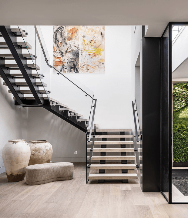 A bright contemporary interior stair hall featuring a sculptural open-riser staircase with dark metal stringers, treads, and slim glass balustrades. Pale timber flooring and white walls create an airy backdrop, complemented by an abstract artwork, two large textured ceramic vessels, and a rounded upholstered bench. A tall dark-framed glazed opening at right reveals a planted vertical garden and exterior paving.