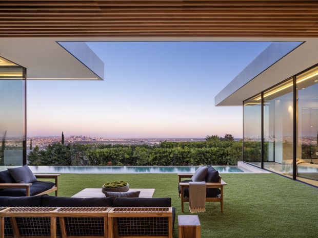 A sophisticated indoor-outdoor living space opening onto a landscaped lawn and narrow infinity-edge pool, with expansive frameless glazing and dramatic cantilevered flat-roof forms. Warm timber slatted soffits frame a broad city skyline and sunset view. The space includes a large outdoor lounge arrangement with timber-framed seating, dark cushions, a low coffee table, and a fire bowl, creating a refined contemporary setting.