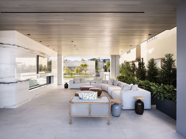A spacious, contemporary covered outdoor living area with a large modular sectional sofa, lounge chairs, coffee tables and decorative stools arranged on a smooth concrete floor. The space has a warm timber-lined ceiling with recessed linear lighting, full-height glazing, a dramatic veined marble-clad fireplace or feature wall, and integrated planter boxes filled with greenery. The open end looks toward a landscaped courtyard and modern residential structures, creating a bright, refined indoor-outdoor setting.