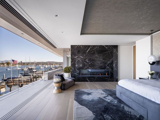 Luxurious contemporary bedroom with floor-to-ceiling glazing overlooking a marina. The room features a large upholstered bed, pale timber flooring, a dark patterned area rug, a dramatic black marble fireplace feature wall, textured concrete ceiling, built-in linear air-conditioning grille, and a sculptural pendant light. An outdoor terrace and waterfront marina are visible beyond the glazing.