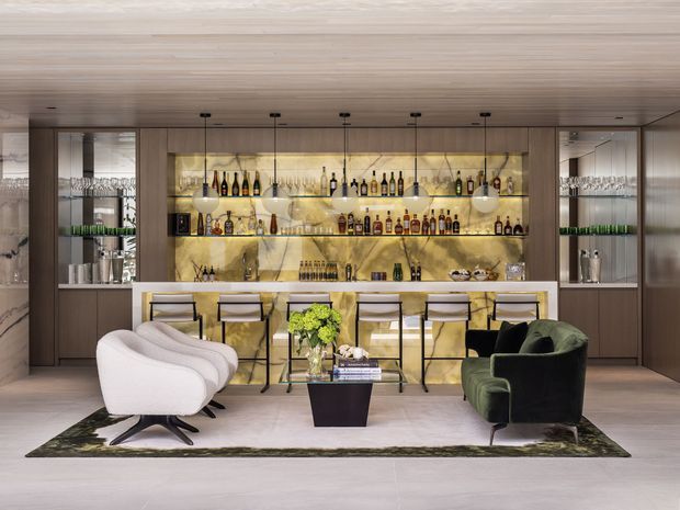 Sophisticated home bar and lounge with a full-width illuminated bottle display, integrated cabinetry, marble-effect bar front, pendant lighting, and seating for four. A sculptural white lounge chair, deep green armchair, coffee table, and area rug create an elegant entertaining space. The room uses warm timber surfaces, dark joinery, and softly lit amber tones.