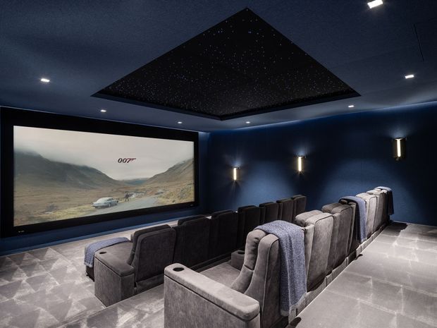 A dedicated home cinema with two rows of large upholstered reclining theater seats facing a wide projection screen. The room has deep navy walls, dark carpeting, concealed and wall-mounted lighting, and a dramatic recessed star-field ceiling feature, creating an immersive cinematic atmosphere.