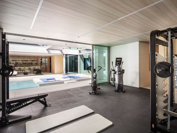 A bright, modern home gym with black rubber flooring, exercise equipment including a squat rack, rowing machine and cardio machine, and light timber-lined ceiling panels. Full-height glazing opens views toward an adjacent courtyard and swimming pool, creating a spacious indoor-outdoor connection.