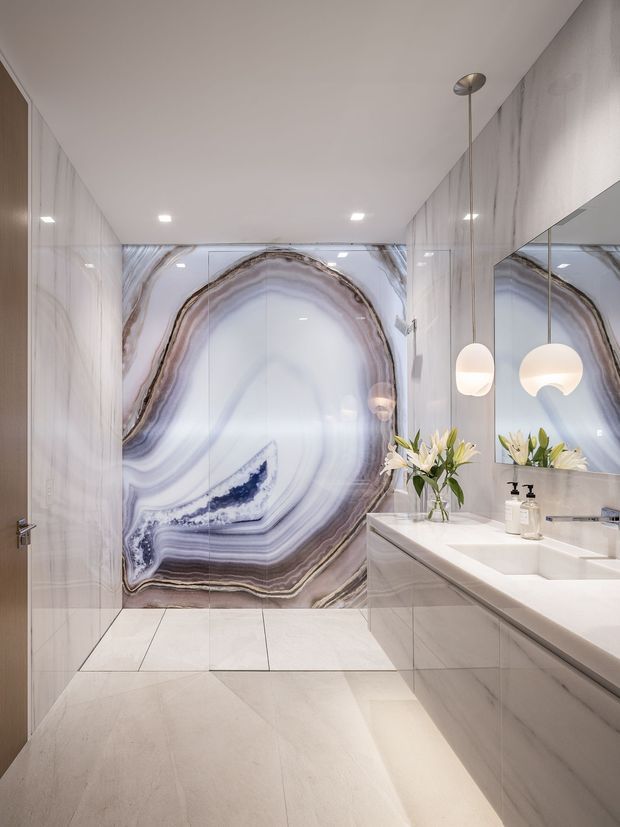 Contemporary luxury bathroom with a walk-in shower enclosed by clear glass, dramatic bookmatched agate-like stone feature wall, large-format pale stone wall and floor tiles, and a long floating double vanity with integrated basins. A large mirror, spherical pendant lights, recessed ceiling lights, and understated botanical styling complete the bright, refined space.