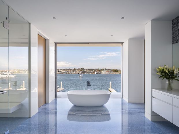 A bright, luxurious bathroom with a freestanding oval bathtub positioned in front of expansive floor-to-ceiling glazing overlooking a waterfront and marina. The room features a highly reflective blue-grey stone or terrazzo floor, crisp white walls, integrated white vanity cabinetry, mirrored surfaces, recessed ceiling lights, and warm timber door detailing.