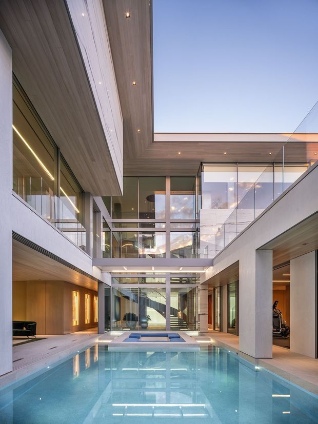 A dramatic contemporary multi-level courtyard centered around a long rectangular reflecting or swimming pool. The space features extensive floor-to-ceiling glazing, exposed structural framing, warm timber soffits and wall cladding, pale rendered or concrete walls, glass balustrades, and recessed linear lighting. Open-air sections reveal a clear sky, while interior rooms and circulation spaces are visible around the courtyard. The composition emphasizes vertical scale, layered terraces, strong geometry, and reflections in the pool.