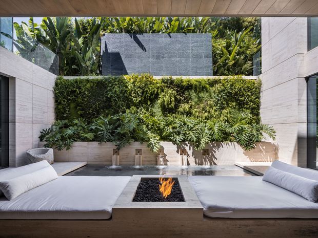 A luxurious contemporary outdoor courtyard lounge framed by pale stone walls and timber soffit, with two low white upholstered daybeds facing a central built-in fire pit. A lush vertical garden and tropical planting create a green feature wall, with a dark stone screen and shallow water feature adding contrast. The space is bright, private, and resort-like.