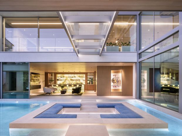 A sophisticated multi-level contemporary interior centered on an indoor courtyard pool. The lower level features a lounge and illuminated built-in bar with timber wall panelling, while the upper level has extensive glass balustrades and a large illuminated architectural ceiling feature. The pool includes a raised geometric sunken seating platform upholstered in blue, surrounded by pale stone or concrete paving. The space is bright, polished, and strongly connected through glass walls and voids.