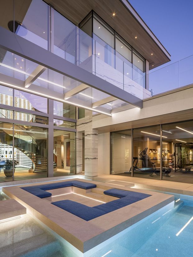 A sophisticated multi-level courtyard featuring a central swimming pool, built-in blue seating, extensive floor-to-ceiling glazing, glass balustrades, and a glazed upper-level bridge. Warm timber soffits, pale stone or concrete surfaces, and illuminated interiors create a refined contemporary architectural setting. A home gym is visible through the glazing, with a sculptural spiral staircase and open living areas beyond.