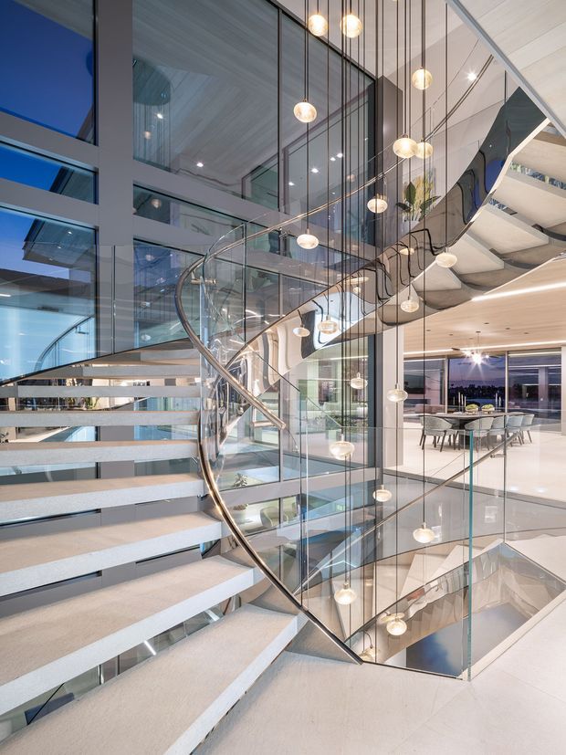 A dramatic contemporary multi-level interior centred on an open stairway with pale stone or concrete treads, clear glass balustrades, and a polished curved metal handrail. A tall glazed atrium rises through the space, with numerous spherical pendant lights suspended at different heights. Dining and lounge areas are visible on the adjacent level, along with extensive glazing, light timber ceiling panels, and crisp white finishes.