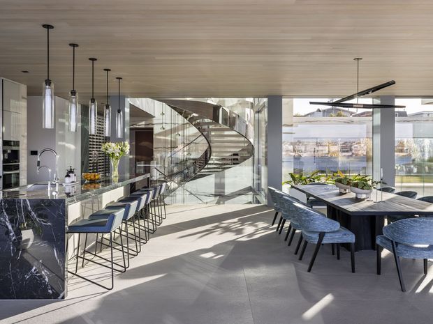 A bright, open-plan contemporary kitchen and dining area with a long dark marble island, integrated sink, blue upholstered bar stools, and slim pendant lights. A sculptural curved staircase with glass balustrades forms a prominent architectural feature behind the kitchen, while floor-to-ceiling glazing overlooks a landscaped waterfront or garden setting. The space has a warm timber ceiling, pale flooring, dark cabinetry and abundant natural light.